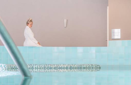 Eine blonde Frau sitzt am Beckenrand des Hotelpools bereit für die Wassergymnastik im Ostseeurlaub im Rugard Thermal Strandhotel.