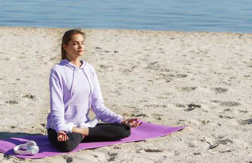 Eine junge Frau in violetten Sportklamotten sitzt am Strand und macht Yoga im Ostseeurlaub im Rugard Thermal Strandhotel.
