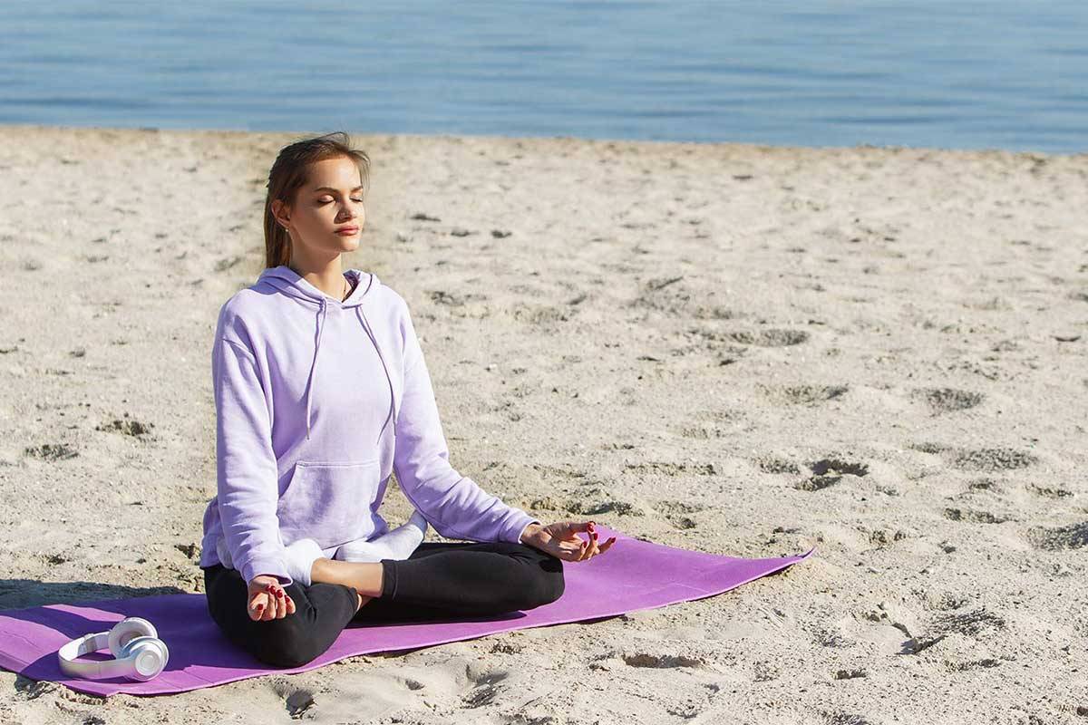 Eine junge Frau in violetten Sportklamotten sitzt am Strand und macht Yoga im Ostseeurlaub im Rugard Thermal Strandhotel.