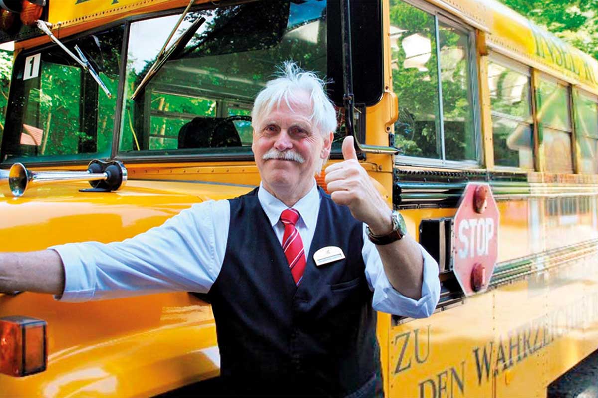 Ein Mann in Uniform lächelt in die Kamera und steht vor einem gelben Schulbus bereit für einen Ausflug des Rugard Thermal Strandhotels.