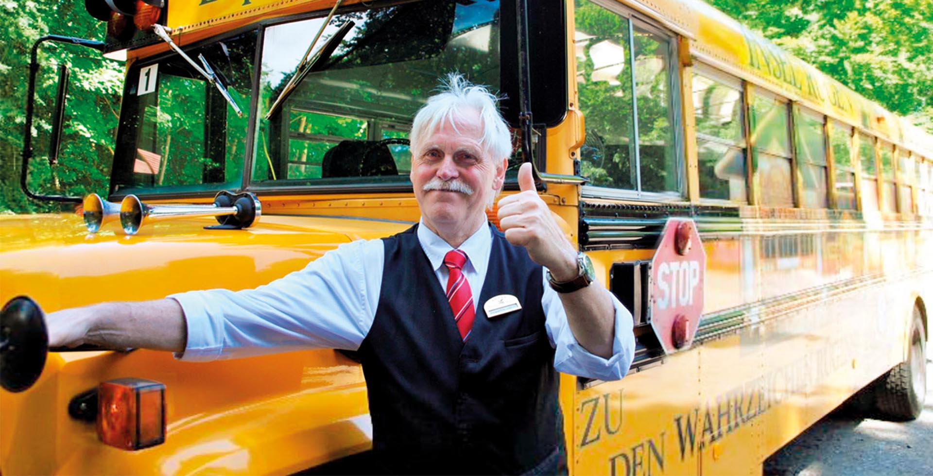 Grinsender Mann hält den Daumen hoch vor einem gelben Schulbus.