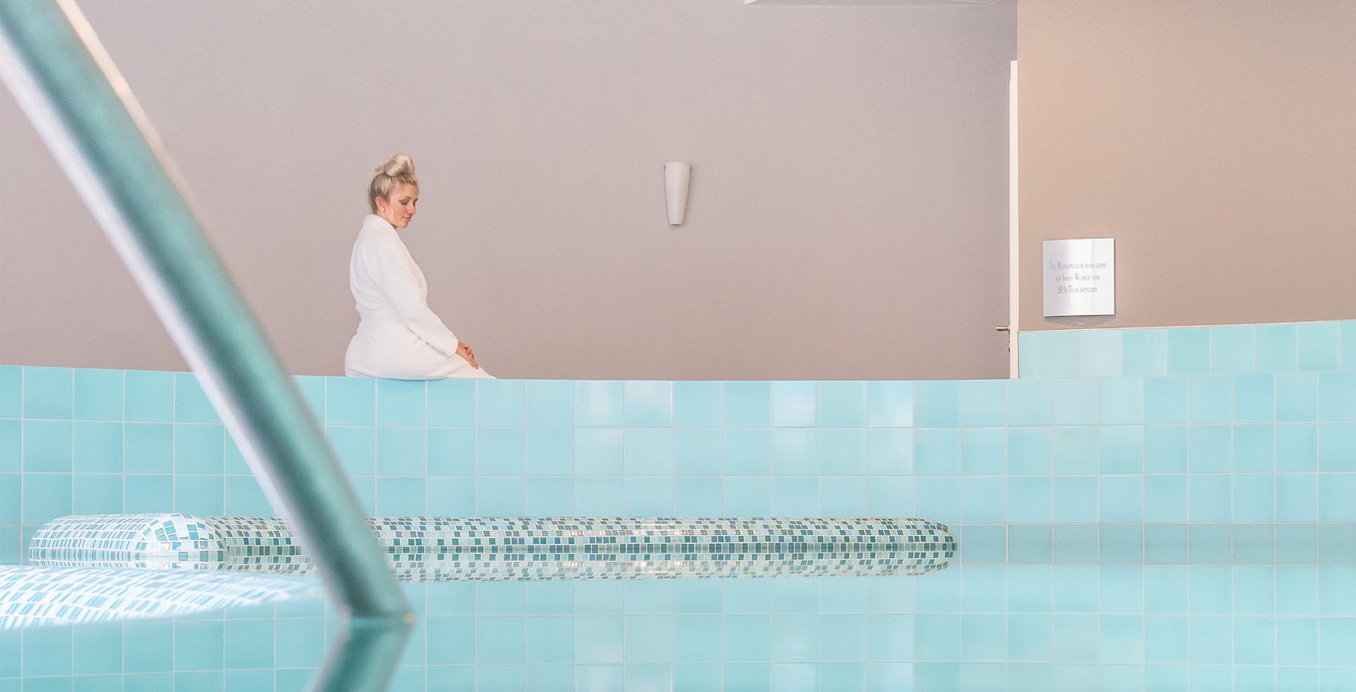 Frau im Bademantel sitzt am Beckenrand eines Indoor Pools.
