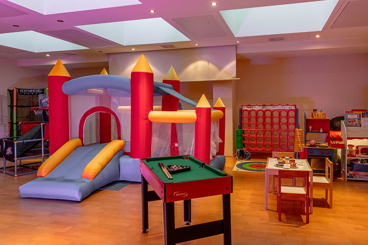 Bunter Kinderspielraum mit Hüpfburg, Kinderküche, kleinem Billardtisch, Tisch und Stühlen für kreative Beschäftigungen und vielen weiteren Utensilien für kinderfreundliche Aktivitäten.