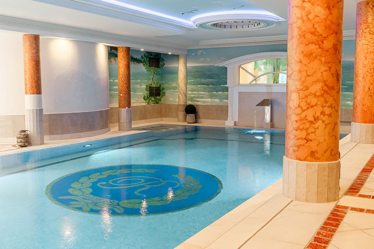 Blaues Schwimmbecken mit dunkelblauem Logo auf dem Boden und Stufen mit weiß-kupferfarbenen Säulen. 2 weiße Liegestühle stehen neben dem Becken. Am Ende des Beckens befindet sich ein von weißen Säulen umrahmter Spiegel.