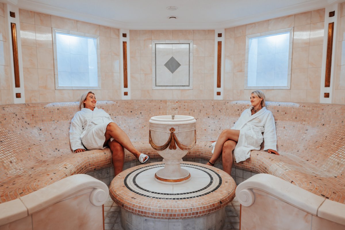 Zwei Frauen sitzen im Caldarium des Rugard Thermal Strandhotels auf beheizten Steinbänken mit Mosaikverkleidung.