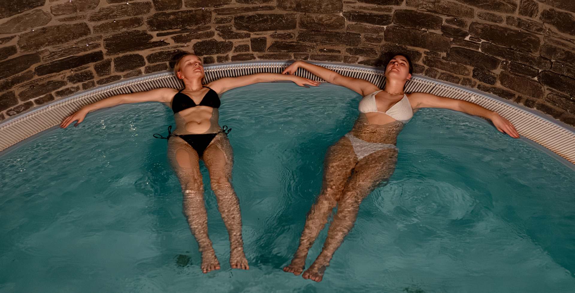 Zwei Frauen entspannen nebeneinander im warmen Wellness-Pool mit Natursteinwand m Rugard Thermal Standhotel auf Rügen.